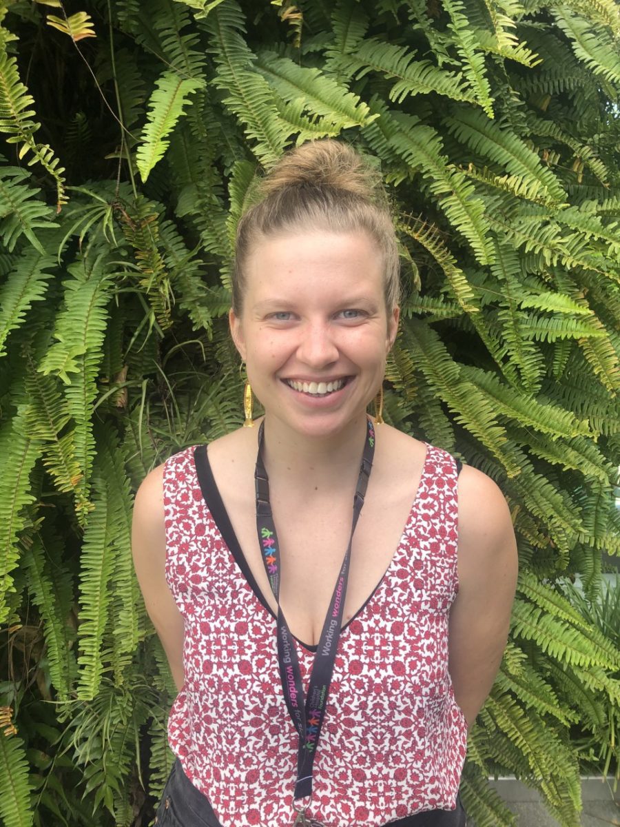 Erin, a music therapist at the Queensland Children's Hospital, wears a red and white patterned top in front of green foliage.
